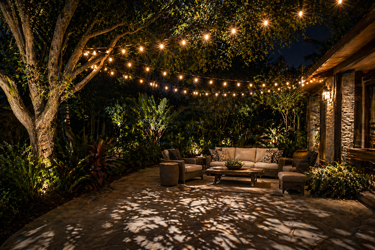 A beautifully lit outdoor living space featuring layered string lights and landscape uplighting, illustrating how professional backyard lighting design creates lasting ambiance and functionality